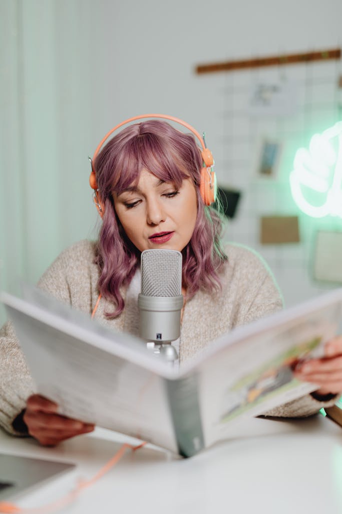 Woman podcasting indoors, reading from a book with microphone and headphones.