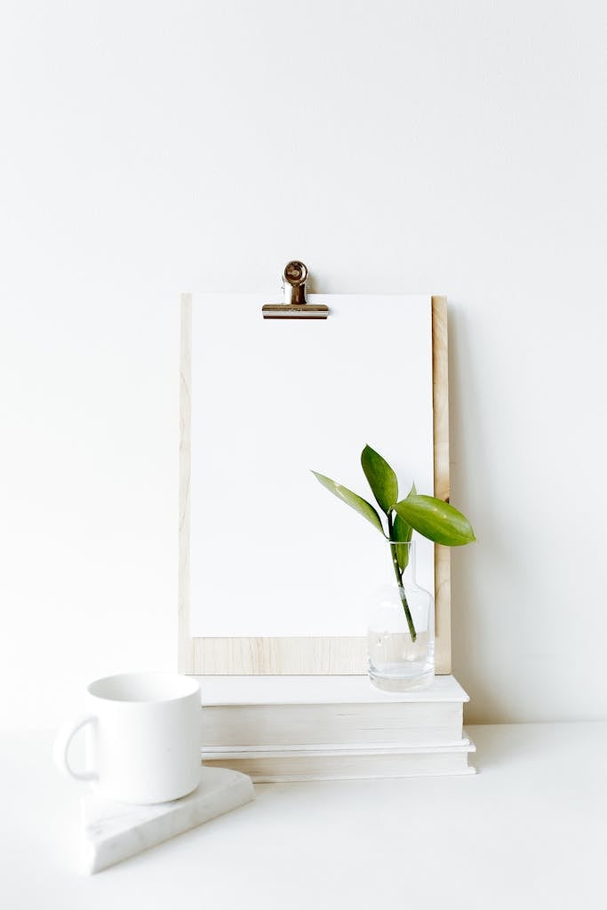 Composition of clipboard with empty sheet placed on stacked books near white mug and green plant leaf